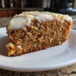 Close-up of a moist slice of carrot cake recipe topped with thick cream cheese frosting, showing visible shredded carrots and walnuts.