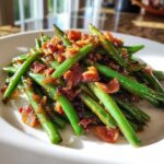 A close-up of bright green beans tossed with crispy bacon bits served on a white plate.