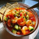 A close-up, vibrant photo of Mexican shrimp cocktail served in a clear glass bowl with shrimp, cucumber, and tomato in a red sauce.