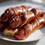 A close-up stack of freshly baked pretzel dogs with a dark, shiny crust and coarse salt crystals.
