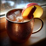 A condensation-covered copper mug filled with an apple cider Moscow mule, garnished with an apple slice and cinnamon stick.