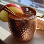 A condensation-covered copper mug filled with an apple cider Moscow mule, garnished with an apple slice and cinnamon sticks.