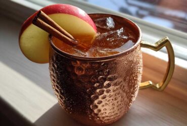 A condensation-covered copper mug filled with an apple cider Moscow mule, garnished with an apple slice and cinnamon sticks.