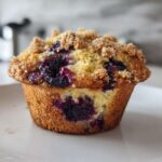 Close-up of a golden brown blueberry muffin topped with crunchy streusel, showcasing burst blueberries.