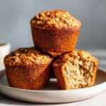A stack of three golden-brown banana oatmeal muffins on a plate, with one muffin broken open to show the soft interior.