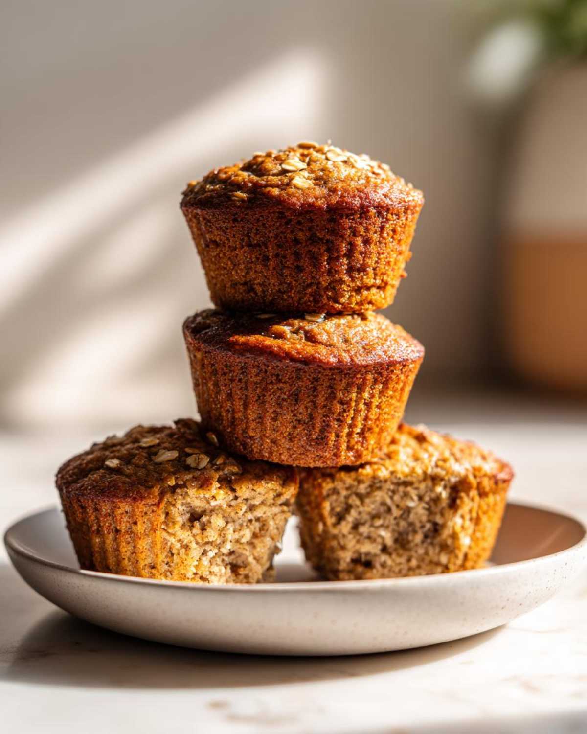 A stack of three golden brown banana oatmeal muffins, one broken open to show the texture.