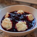 A white bowl filled with warm Blackberry Dumplings swimming in a rich, dark purple blackberry sauce.