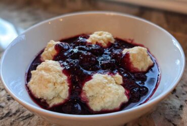 A white bowl filled with warm Blackberry Dumplings swimming in a rich, dark purple blackberry sauce.