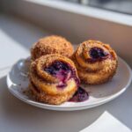 Three golden-brown Blueberry Pie Bombs coated in cinnamon sugar, one showing gooey blueberry filling spilling out.