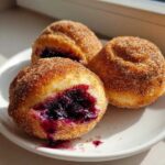 Three golden, cinnamon-sugar coated Blueberry Pie Bombs, one cut open revealing rich blueberry filling, sitting on a white plate.