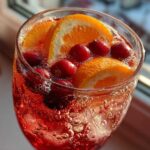 Close-up of a glass filled with bubbly, red non-alcoholic Christmas punch garnished with orange slices and fresh cranberries.