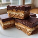Three stacked and arranged squares of rich buckeye graham crackers bars with chocolate topping and caramel layer.