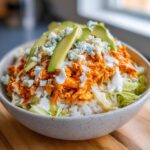A close-up of a vibrant buffalo chicken bowl featuring shredded orange buffalo chicken over rice and lettuce, topped with avocado slices, blue cheese crumbles, and ranch dressing.