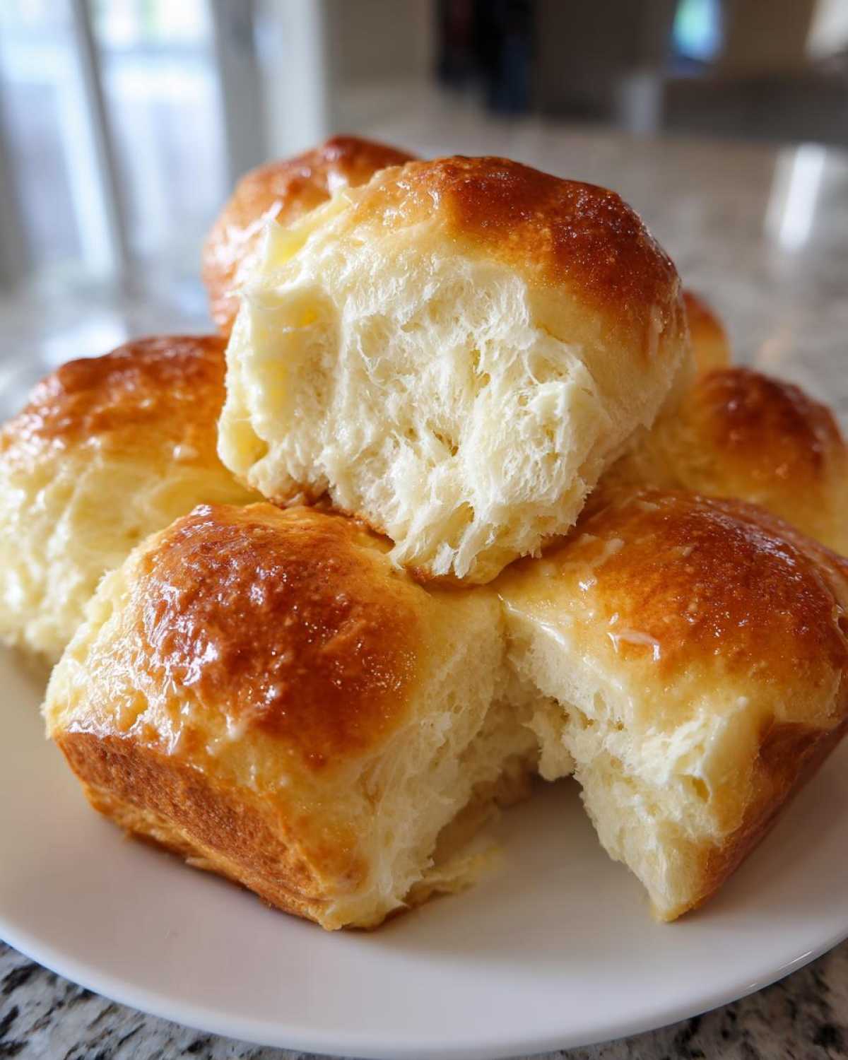 Close-up of several fluffy, golden-brown buttery rolls stacked on a white plate, one torn open to show the soft interior.