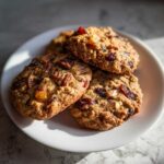 A close-up of three chewy fruitcake cookies loaded with dried fruit and nuts on a white plate.