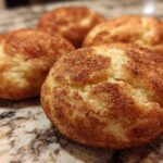 Close-up of four freshly baked, chewy snickerdoodle cookies heavily coated in cinnamon sugar resting on a granite countertop.
