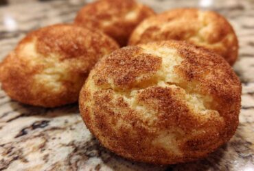 Close-up of four freshly baked, chewy snickerdoodle cookies heavily coated in cinnamon sugar resting on a granite countertop.