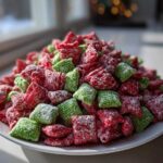 A mound of bright red and green Christmas puppy chow covered in powdered sugar, sitting on a white plate near a window.