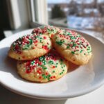 Four soft, round christmas sprinkle sugar cookies topped with red, green, and white nonpareils, sitting on a white plate.