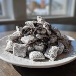 A generous mound of cookies and cream puppy chow coated in powdered sugar and dark cookie crumbs, served on a white plate.