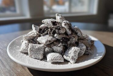 A generous mound of cookies and cream puppy chow coated in powdered sugar and dark cookie crumbs, served on a white plate.