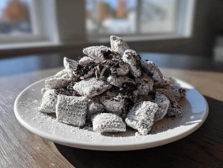 A generous mound of cookies and cream puppy chow coated in powdered sugar and dark cookie crumbs, served on a white plate.