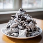 A large mound of cookies and cream puppy chow coated in powdered sugar and dark cookie pieces, served on a white plate.