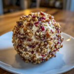 A close-up of a perfectly formed cranberry pecan cheese ball coated in chopped nuts and dried cranberries, resting on a white plate.