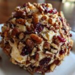 A close-up view of a perfectly formed cranberry pecan cheese ball coated in chopped pecans and dried cranberries.