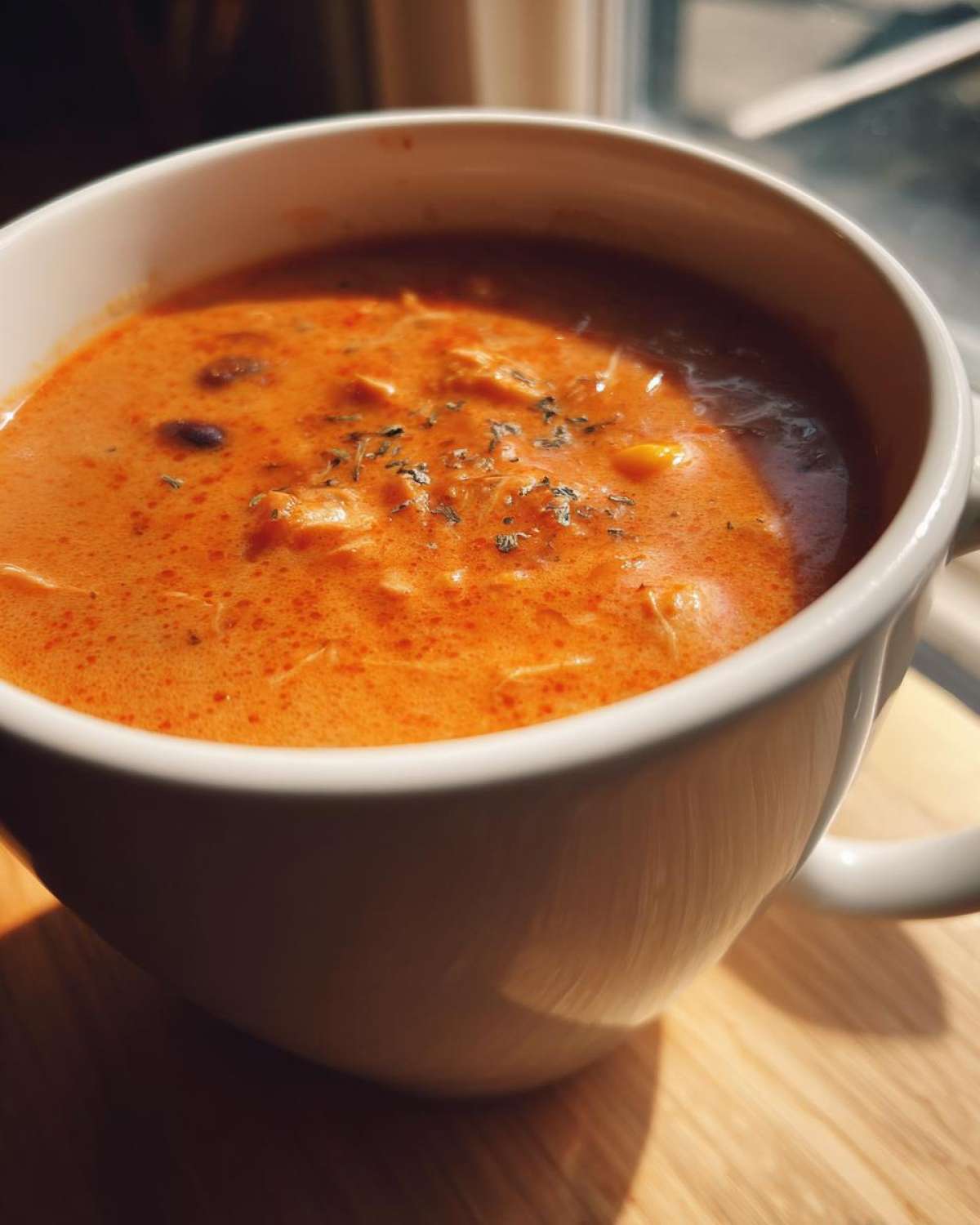 Close-up of creamy, orange-hued chicken taco soup garnished with herbs in a white mug.