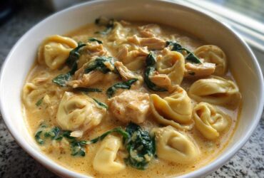 A close-up shot of a white bowl filled with rich, creamy chicken tortellini soup featuring spinach and chunks of chicken.