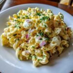 A close-up of creamy deviled egg pasta salad mixed with chopped red onion and parsley on a white plate.