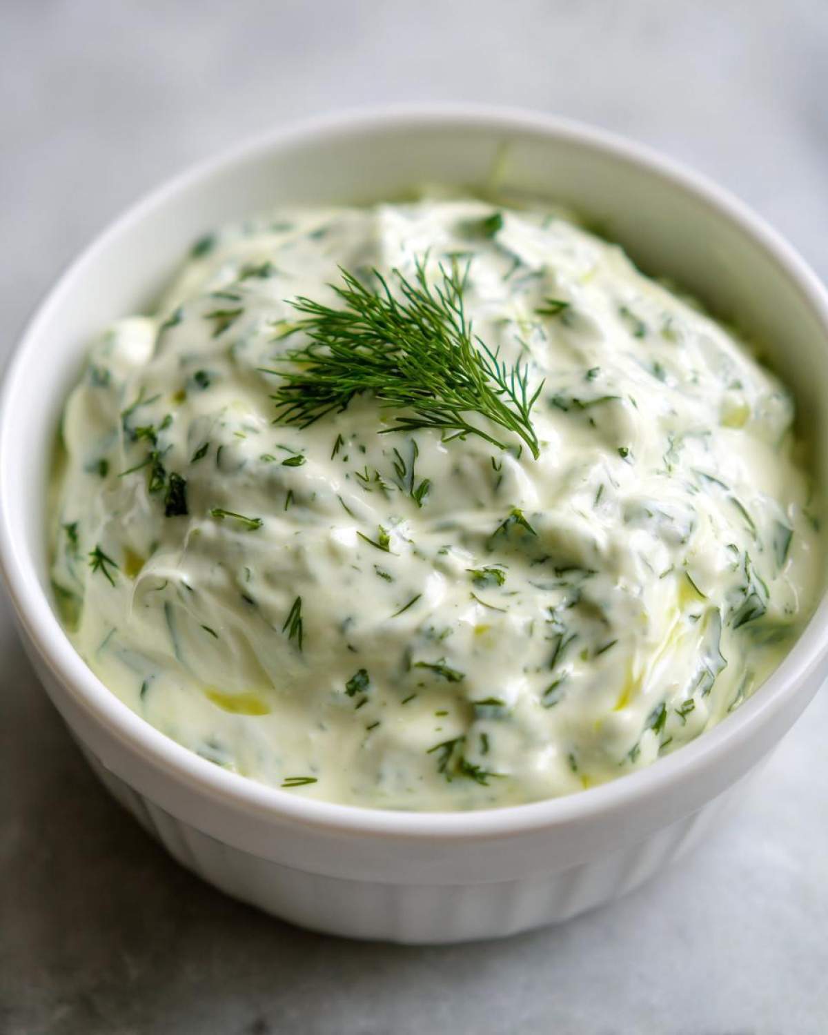 Close-up of creamy, herb-filled dill dip garnished with a fresh sprig of dill in a white bowl.