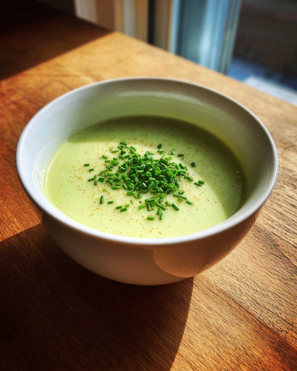 A bowl of creamy, light green leek soup garnished generously with chopped chives and a sprinkle of spice.