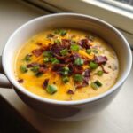 A close-up of creamy loaded baked potato soup topped generously with shredded cheddar cheese, bacon bits, and green onions.