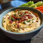 A bowl of creamy Million Dollar Dip topped generously with crumbled bacon and fresh chopped chives, served with vegetable sticks.