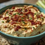 A close-up of creamy million dollar dip served in a teal bowl, topped generously with crumbled bacon and chopped green onions.