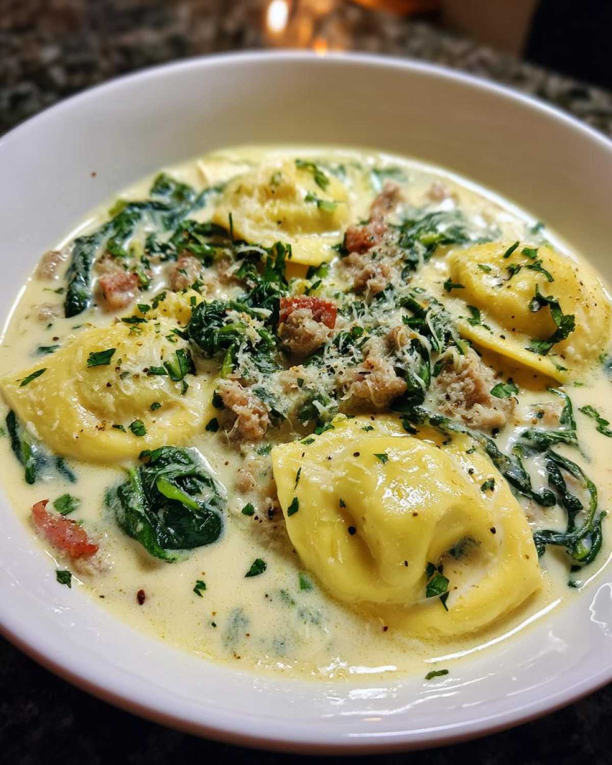 A close-up of a bowl of creamy ravioli soup featuring large pasta pockets, wilted spinach, sausage crumbles, and grated Parmesan cheese.