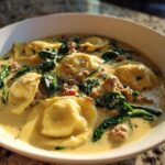 A close-up of a white bowl filled with creamy ravioli soup featuring cheese ravioli, crumbled sausage, and wilted spinach.