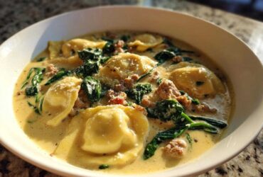 A close-up of a white bowl filled with creamy ravioli soup featuring cheese ravioli, crumbled sausage, and wilted spinach.