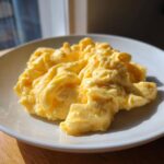 Close-up of fluffy, creamy scrambled eggs served on a white plate, highlighted by natural sunlight.