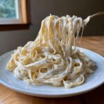 A fork lifts a large tangle of fettuccine heavily coated in creamy vegan alfredo sauce from a white plate.