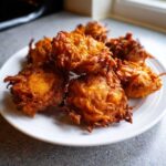 A stack of golden brown, crispy Amish Onion Fritters piled high on a white plate.