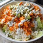 A close-up of a white bowl filled with a buffalo chicken bowl featuring shredded orange buffalo chicken, white rice, lettuce, avocado, and blue cheese crumbles.