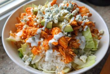 A close-up of a white bowl filled with a buffalo chicken bowl featuring shredded orange buffalo chicken, white rice, lettuce, avocado, and blue cheese crumbles.