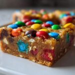 A close-up of a thick, chewy square of christmas cookie bars topped with colorful candy-coated chocolates.