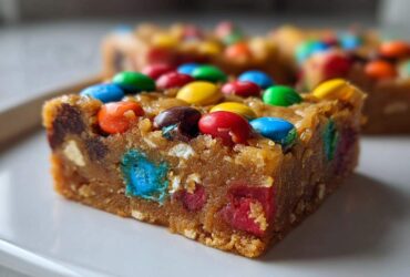 A close-up of a thick, chewy square of christmas cookie bars topped with colorful candy-coated chocolates.