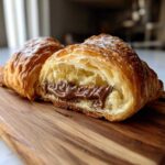 A flaky, golden-brown chocolate croissant cut in half, revealing a rich, melted chocolate filling inside.