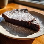 A rich, dark slice of flourless chocolate torte dusted heavily with powdered sugar on a light plate.