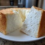A tall, fluffy slice of angel food cake with a golden crust sitting next to the rest of the cake on a white plate.
